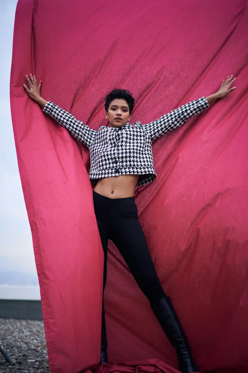 Kazayağı ceketli model Taahira kolları iki yana açık, kırmızı kumaşın önünde siyah çizmelerle, çatı katında editoryal moda fotoğrafı, Ankara moda fotoğrafçısı Şems Erik.